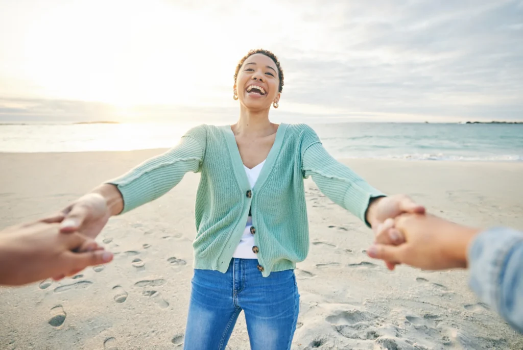 Happy woman reaching out and holding hands with others on a beach during sunset, symbolizing support and connection in recovery.